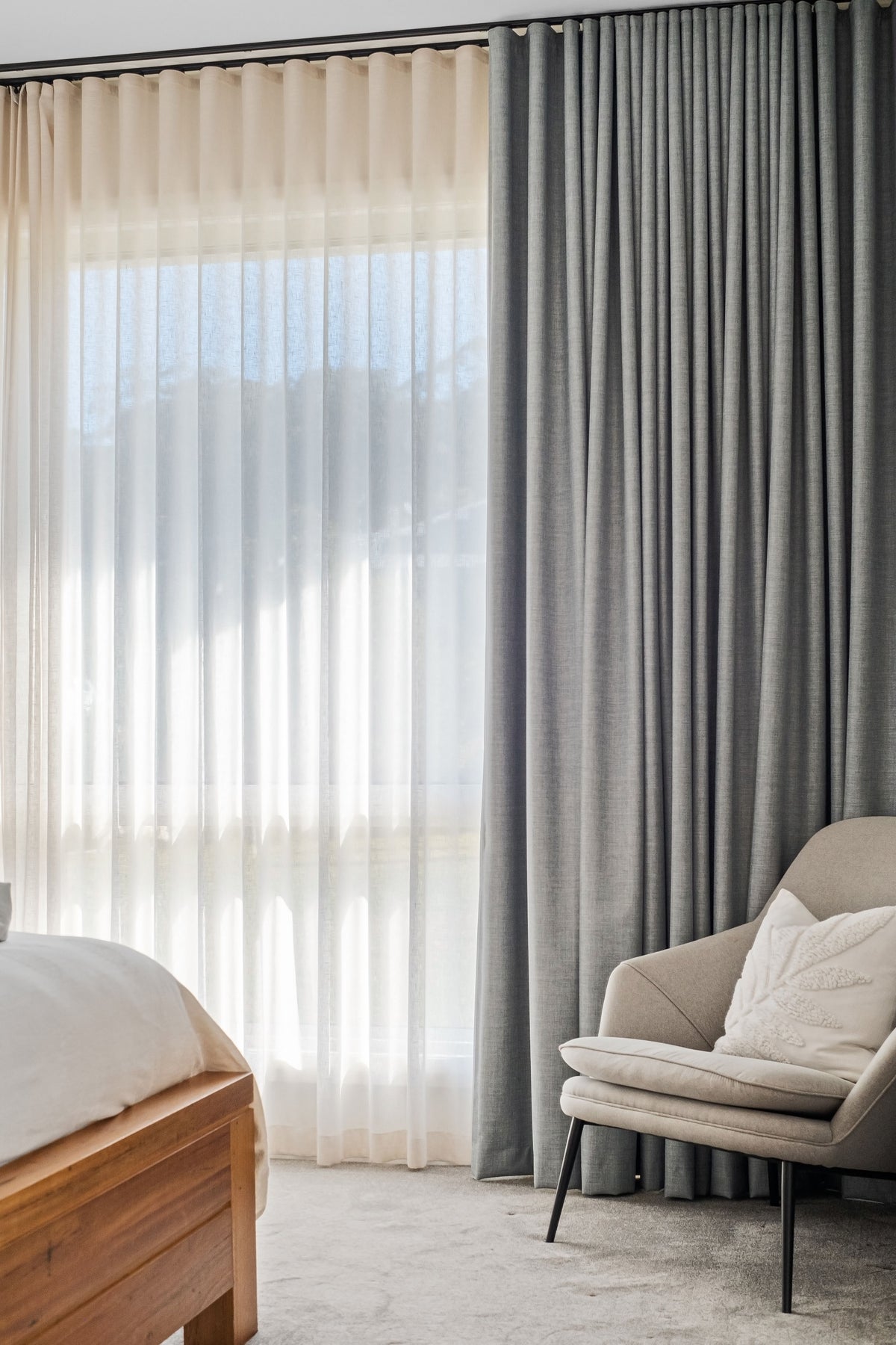 Soft grey curtains layered with sheers beside a bed and an upholstered chair